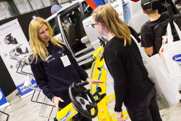 A woman is demonstrating a yellow accessible vehicle steering wheel to a visitor at the ATSA Sydney Expo 2019