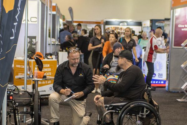 A man kneeling down talking to a wheelchair user on the expo floor - ATSA Brisbane image gallery