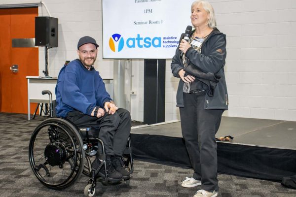 A woman holding a microphone with a male wheelchair user showcasing adaptive clothing items during ATSA Expo Fashion gallery